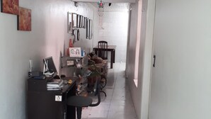 Ein schmaler Korridor mit einem Schreibtisch, einem Bürostuhl und einem weißen Hocker. An der Wand befinden sich gerahmte Bilder und ein kleiner Tisch mit Gegenständen darauf.
