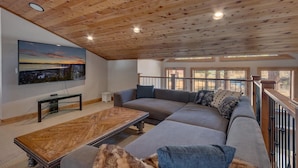 Ein geräumiges Wohnzimmer mit Holzbalkendecke, einem großen Sofa, einem Couchtisch und einem an der Wand befestigten Fernseher.