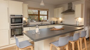 Une cuisine moderne avec des meubles blancs, un grand îlot central doté de tabourets, des appareils en acier inoxydable et une fenêtre laissant entrer la lumière naturelle.