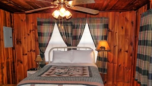 Dormitorio de cabaña de madera con cama, ventilador de techo, cortinas de cuadros y lámpara.