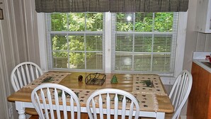 Un comedor con una mesa de madera y sillas blancas, un valance a cuadros sobre la ventana, y vistas a zonas verdes al exterior.