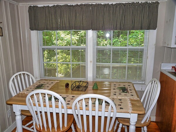 A dining area with a wooden table and white chairs, a checkered valance above the window, and a view of greenery outside. - Cozy Cabin on the Creek-Creek side- Secluded - Charcoal Grill (Bryson City)
