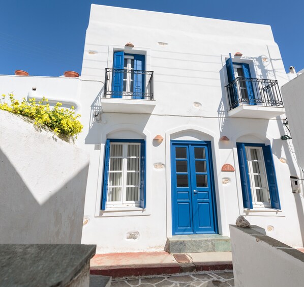 Ein weißes Gebäude mit blauen Türen und Fenstern, einem kleinen Balkon und einer Steinmauer mit gelben Blumen.