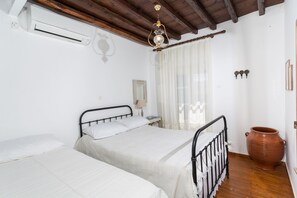 Une chambre avec deux lits, un plafond en bois, un climatiseur, un grand pot en terre cuite et une fenêtre avec des rideaux légers.