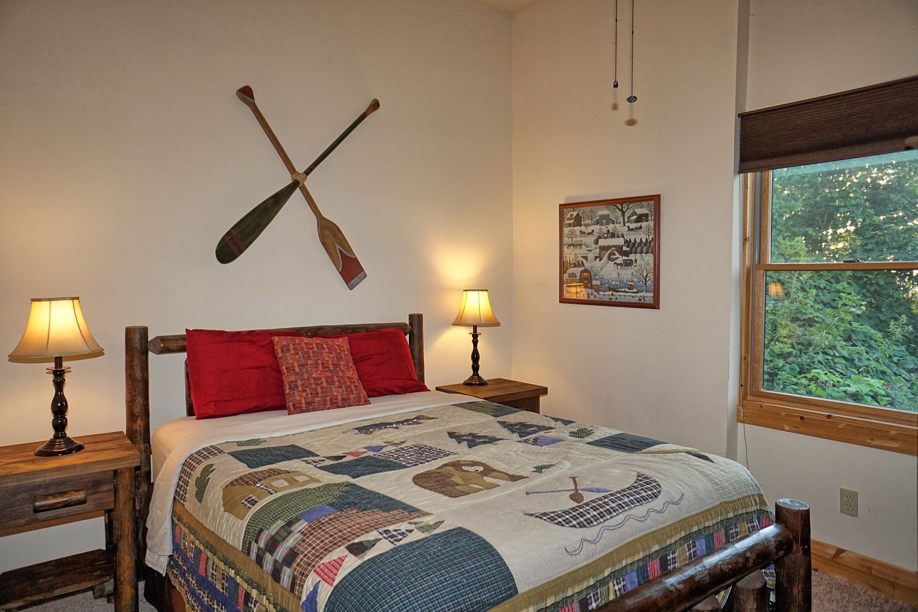 A bedroom with a wooden bed, two bedside tables with lamps, a quilt, a wall-mounted paddle, and a window with a view of greenery.
