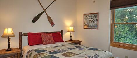 A bedroom with a wooden bed, two bedside tables with lamps, a quilt, a wall-mounted paddle, and a window with a view of greenery.