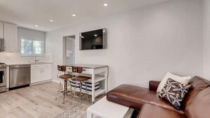 Una cucina moderna con mobili bianchi, una lavastoviglie in acciaio inossidabile e un forno integrato. C'è un'area pranzo con due sgabelli e un tavolo bianco. Nell'area soggiorno si trova un divano in pelle marrone con cuscini decorati.