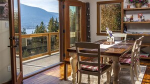 Comedor con mesa y sillas de madera, vista a un lago y montañas a través de amplios ventanales, y estante con objetos decorativos.