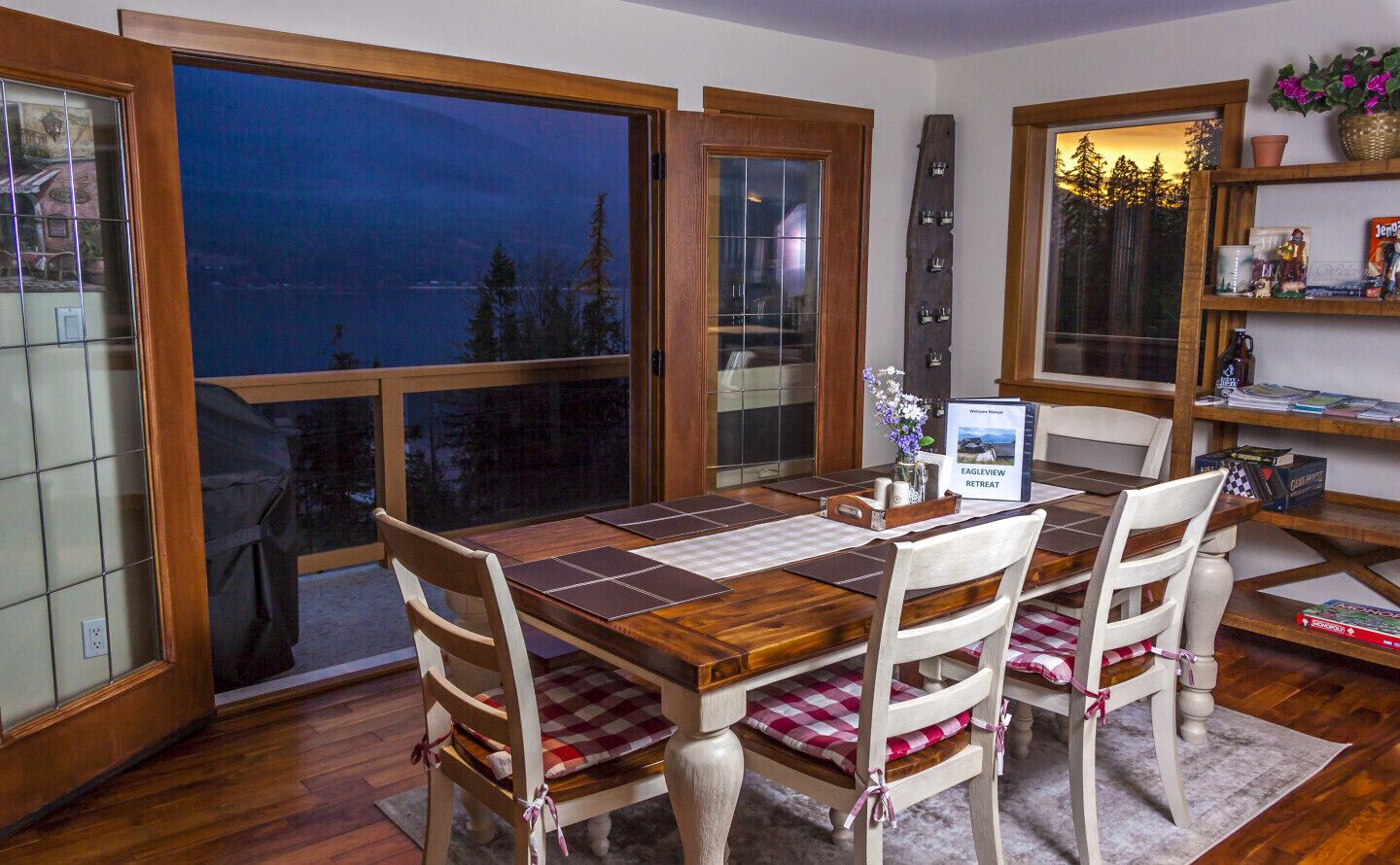 Un coin repas avec une table et des chaises en bois, une vue sur l’extérieur grâce à de grandes fenêtres, et une étagère contenant divers objets.