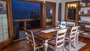 Un comedor con una mesa y sillas de madera, vistas al exterior a través de amplios ventanales, y una estantería con diversos objetos.