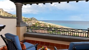 A balcony with two blue chairs, a small table with fruit, and a view of the beach and ocean. - Ocean View Suites to Accommodate Any Party Size On the Beach (Cabo San Lucas)