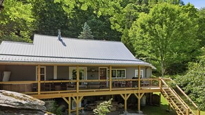 A cabin with a deck, surrounded by trees and rocks. - Alamo River House in Boone, NC: A charming 3BR chalet for 8 by the river! (Boone)