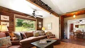 A cozy living room with a brown leather sofa, a coffee table, and a dining area with wooden chairs and a table. - Alamo River House in Boone, NC: A charming 3BR chalet for 8 by the river! (Boone)