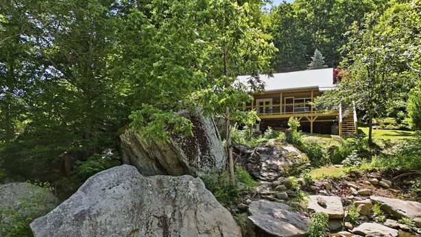 A natural landscape with a stream, large rocks, and a building with a red roof in the background. - Alamo River House in Boone, NC: A charming 3BR chalet for 8 by the river! (Boone)
