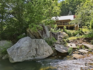 A natural landscape with a stream, large rocks, and a building with a red roof in the background.