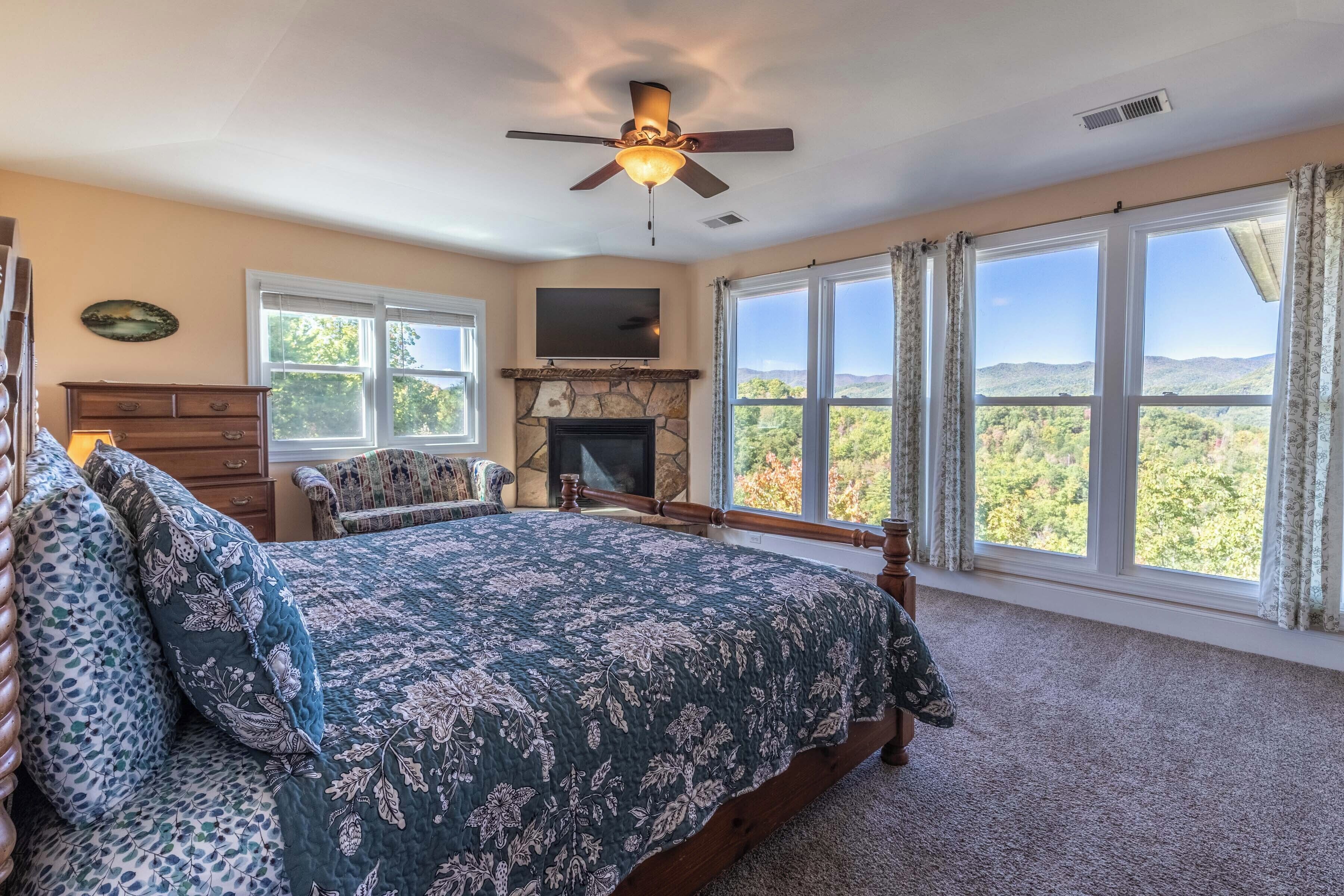 A bedroom with a large bed, a fireplace, a ceiling fan, and a view of the outdoors.