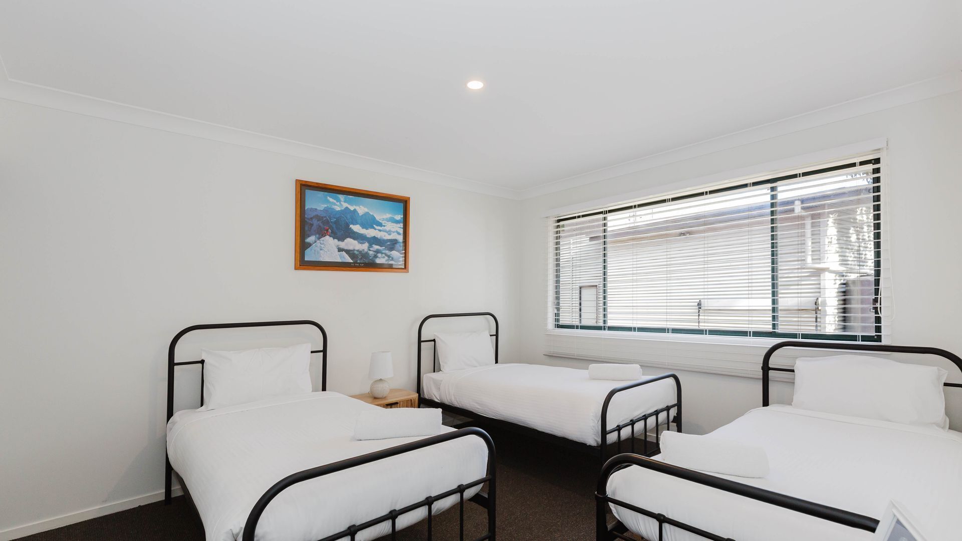 A hotel room with three single beds, a window with blinds, a painting on the wall, and a ceiling light.