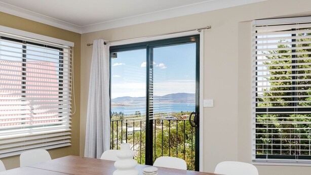 Una habitación con una mesa de comedor, sillas, vistas a un balcón con barandilla a través de un vidrio corredero, y una ventana con persianas.