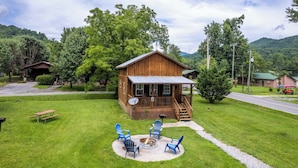 A rustic cabin with a porch, surrounded by a well-maintained lawn and a fire pit area with chairs.