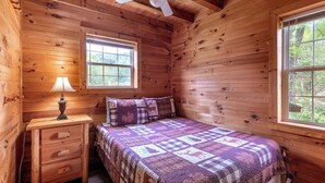 A wooden cabin bedroom with a bed, nightstand, lamp, ceiling fan, and two windows.