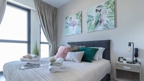 Una cama hecha con cuidado, con un edredón blanco texturizado, un almohadón rosa suave y un almohadón verde. Hay dos cuadros colgados en la pared con flamencos. Se ve una mesita de noche con una lámpara y un reloj.