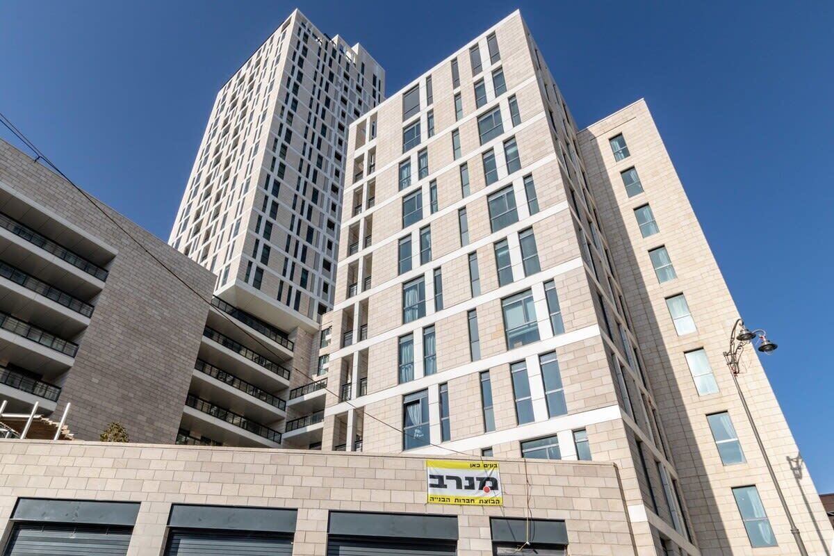 A multi-story building with a modern architectural design, featuring a mix of window patterns and a prominent yellow sign with Hebrew text.