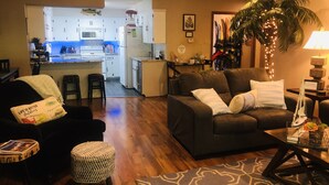 Ein gemütliches Wohnzimmer mit einer dunkelbraunen Couch, einem kleinen runden Hocker und einem Couchtisch. Im Hintergrund ist eine Küche mit weißen Schränken und einer Mikrowelle zu sehen.