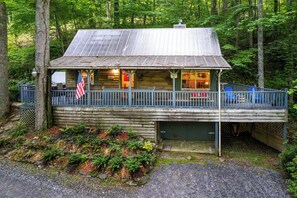 A rustic cabin with a covered porch, a flag, and a gravel driveway.