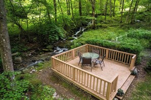 A wooden deck with a glass table and chairs overlooking a forested area with a stream.