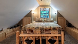 A loft bedroom with a sloped ceiling, a large bed with a wooden frame, a window with a wreath, and a view of trees outside. - Cabin on the Creek-Creek side- Hot tub - Fire Pit - Sleeps 4 (Whittier)