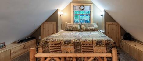 A loft bedroom with a sloped ceiling, a large bed with a wooden frame, a window with a wreath, and a view of trees outside.