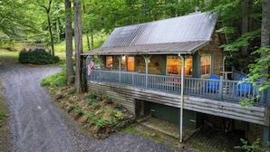 A rustic cabin with a porch, a flag, and a view of the forest. - Cabin on the Creek-Creek side- Hot tub - Fire Pit - Sleeps 4 (Whittier)