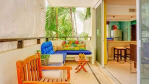 Un balcón con sofá, una silla de madera, una mesita y una cocina visible a través de una puerta abierta.