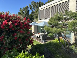 Un espacio moderno para comer al aire libre con una mesa y sillas, rodeado de abundante vegetación y un arbusto rojo con flores.