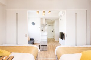 Una habitación de hotel moderna con dos camas, una mesita, un televisor y una kitchenette al fondo.