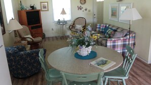 Un salon chaleureux avec une table ronde, des fauteuils verts, un canapé à carreaux et un téléviseur en arrière-plan.