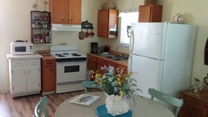 Une cuisine compacte avec des meubles blancs, une cuisinière, un four à micro-ondes et un réfrigérateur. On y trouve une petite table à manger avec des chaises et un vase de fleurs.