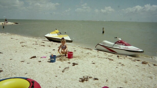 Une plage de sable fin avec un jet ski et un bateau à moteur, un enfant qui joue dans le sable et un ciel dégagé.