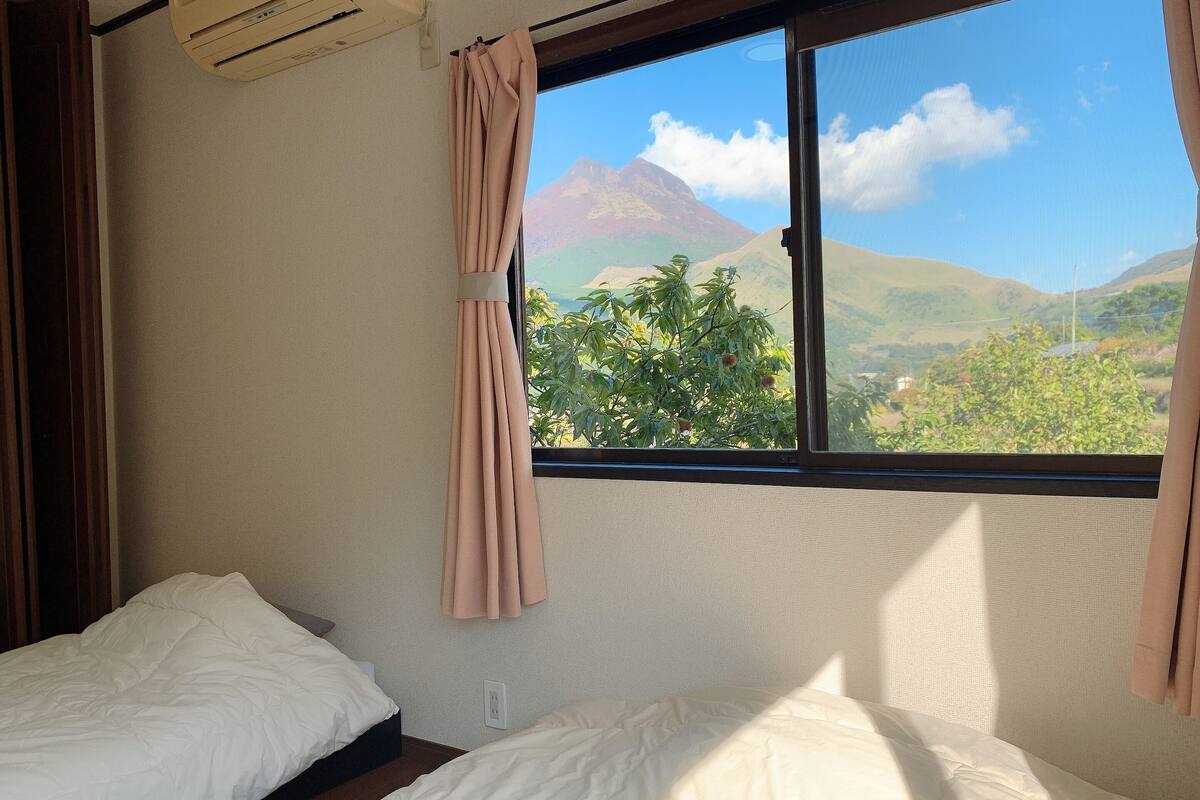A bedroom with two beds, a window with curtains, an air conditioning unit, and a view of a mountain landscape.