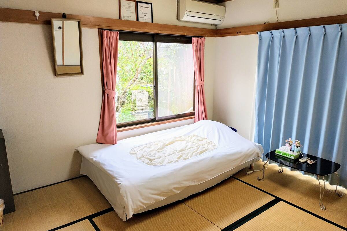 A room with a bed, a window with pink curtains, an air conditioner, and a small table with a decorative item.
