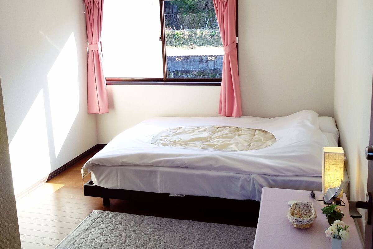 A bedroom with a bed, a bedside table with a lamp, a window with curtains, and an air conditioning unit.