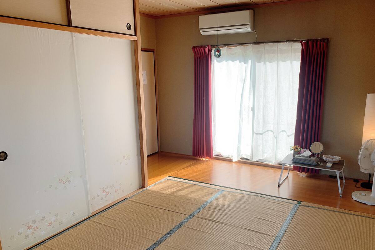 A room with tatami flooring, a sliding door, an air conditioning unit, a table with a fan, and a window with curtains.