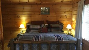 A wooden cabin bedroom with a bed, two bedside lamps, and a painting on the wall.