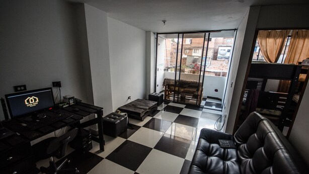 Une pièce avec un sol à carreaux noirs et blancs, un canapé en cuir noir, un bureau avec un ordinateur et un écran, et une porte-fenêtre coulissante donnant sur un balcon.