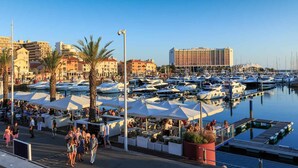 A marina with numerous boats docked, a hotel in the background, and outdoor seating areas with umbrellas.