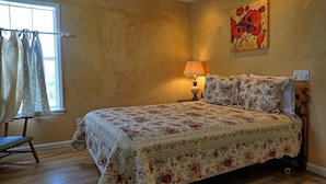 Una camera da letto con una coperta a motivi floreali, un comodino con una lampada, una sedia e una finestra con tende.