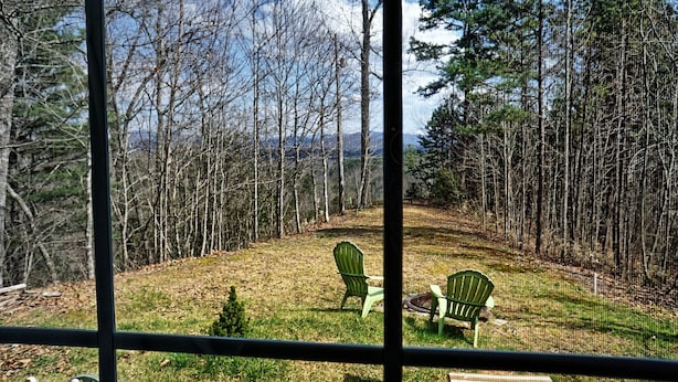 Vista attraverso una finestra su un giardino retrostante con due sedie verdi, un tavolino e una zona erbosa con alberi.