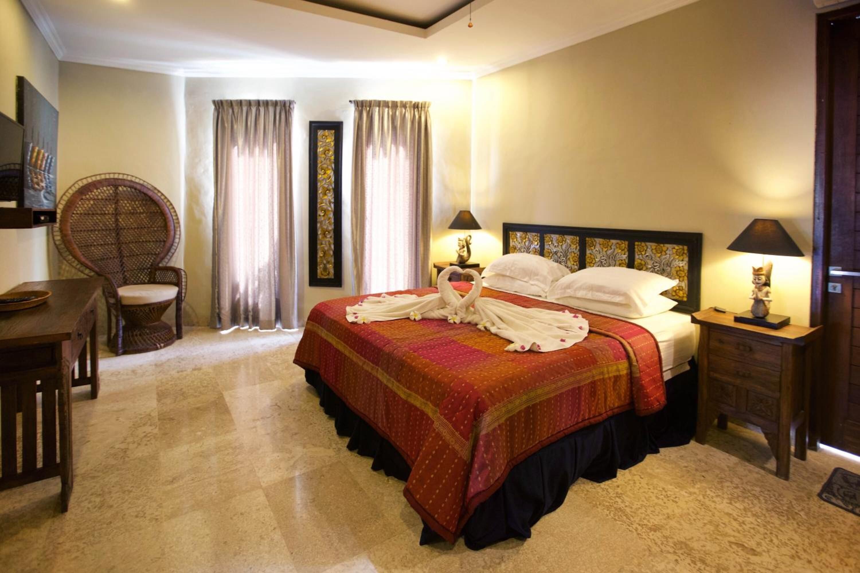 Una habitación de hotel con una cama grande, dos mesitas de noche con lámparas, una silla de madera, una mesa de comedor y cortinas.