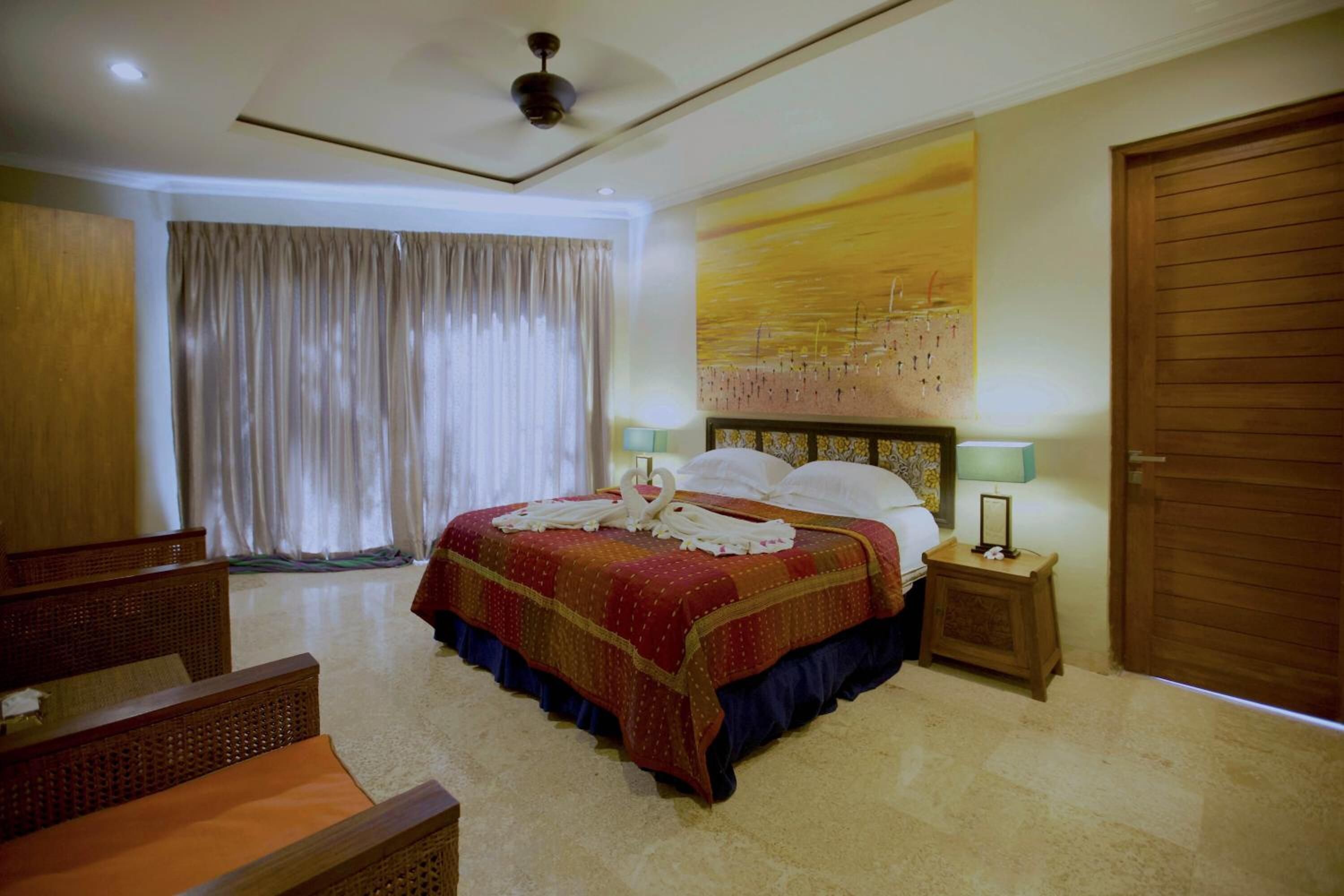 Une chambre d’hôtel avec un grand lit, deux tables de chevet, un ventilateur de plafond, une grande fenêtre avec des rideaux et une porte en bois.