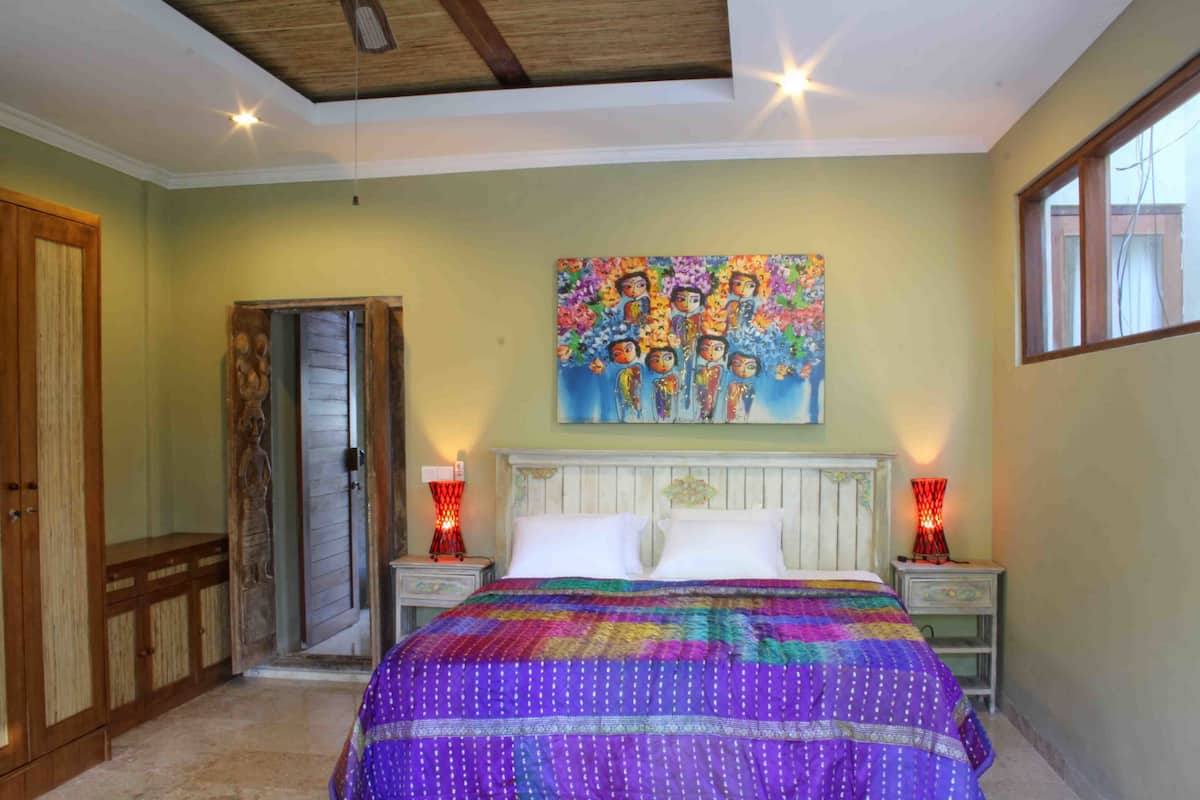 A bedroom with a bed, two bedside tables, a nightstand, a wooden door, and a wall painting.